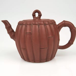 May include: A red clay teapot with a bamboo design. The teapot has a round body and a lid with a small knob on top. The handle is shaped like a bamboo stalk.