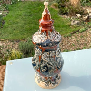 Może przedstawiać: Ozdobny ceramiczny wazon z pokrywką. Wazon ma szczegółowy wzór w odcieniach czerwieni, czerni, kremu i niebieskiego. Wazon ma pękaty kształt z podstawą na cokole i spiczastym wierzchołkiem. Wazon jest wystawiony na szklanym stole.