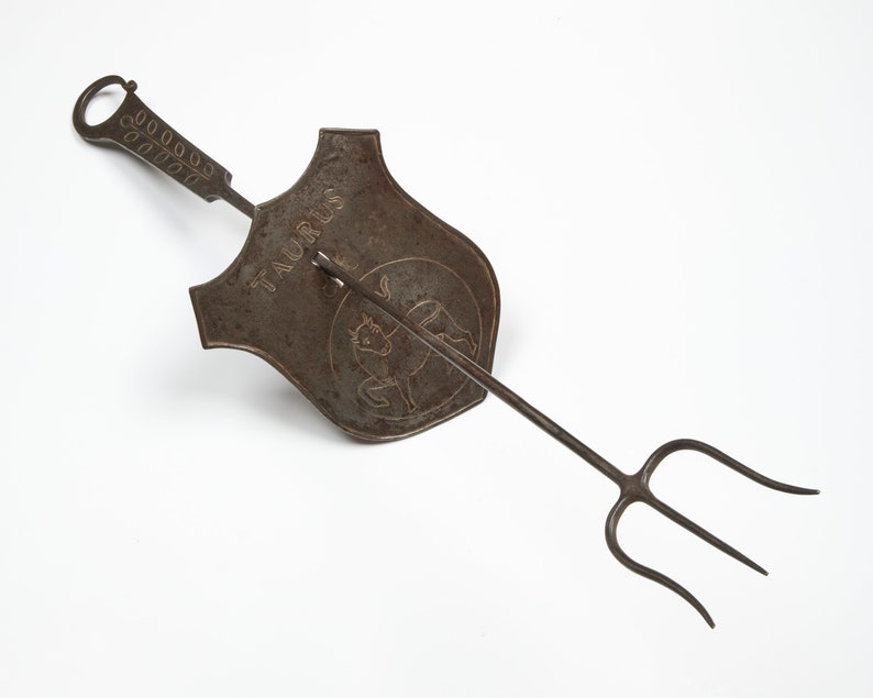 Puede incluir: Una herramienta de chimenea de metal con un mango largo y una horquilla de tres puntas. El mango tiene una pieza decorativa en forma de escudo con la palabra "TAURUS" y un toro grabado.
