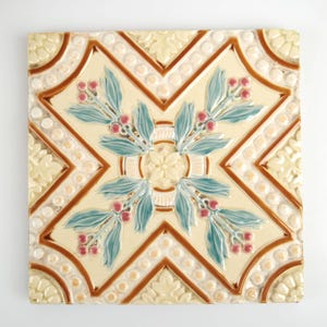 Könnte beinhalten: Quadratische Keramikfliese mit floralem Design. Die Fliese hat einen cremefarbenen Hintergrund mit einem braunen geometrischen Muster. Das Design umfasst türkisfarbene Blätter und rote Beeren. Die Fliese ist etwa 30 cm groß.