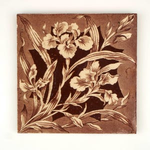 Può includere: Una piastrella di ceramica marrone con un motivo floreale bianco. La piastrella presenta un motivo ripetitivo di fiori di iris e foglie.