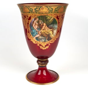 以下が含まれることがあります： 金色のアクセントと男性と女性の絵が描かれた、背の高い赤いガラスの花瓶。花瓶は幅広で丸い底と細い首をしています。