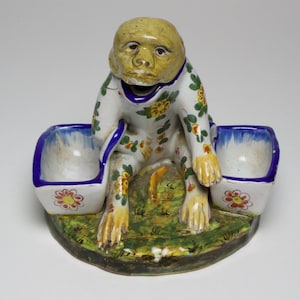 May include: A ceramic monkey figurine with a yellow face and blue and white floral patterned body. The monkey is holding two small white bowls with blue rims.