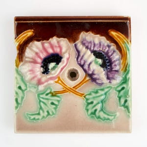 May include: Square ceramic tile with a design of two poppy flowers in pink and purple, with green leaves and stems. The tile has a brown border and a central hole, possibly for a decorative purpose or part of a larger design.