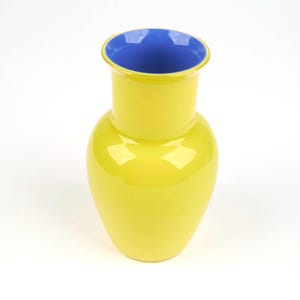 May include: A bright yellow glass vase with a smooth, glossy finish and a contrasting deep blue interior. The vessel features a rounded body that tapers into a cylindrical neck with a flared rim, showcasing a vibrant two-tone colour scheme.