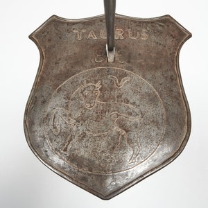 Puede incluir: Una herramienta de fuego de metal con una cabeza en forma de escudo. El escudo tiene un toro grabado en &eacute;l y la palabra "Taurus" escrita encima.