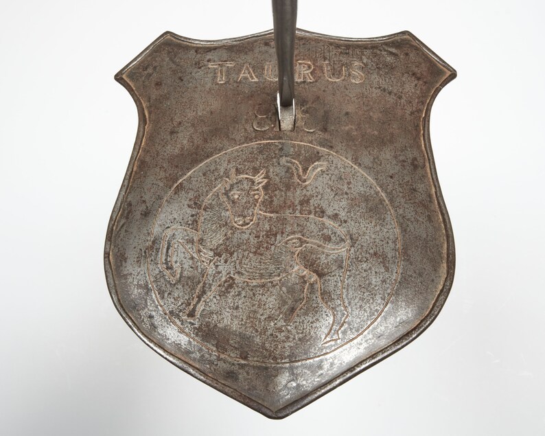 Puede incluir: Una herramienta de fuego de metal con una cabeza en forma de escudo. El escudo tiene un toro grabado en &eacute;l y la palabra "Taurus" escrita encima.