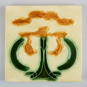 May include: A square ceramic tile featuring a raised design of stylised flowers in shades of brown and green. The flowers are arranged in a symmetrical pattern with a central stem and two curved branches.