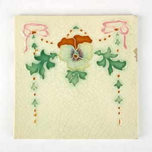 May include: Square ceramic tile with a floral design. The tile features a central flower with a purple center, surrounded by green leaves and accented with pink bows and hanging green and orange details. The background is a cream color with a crackle finish.