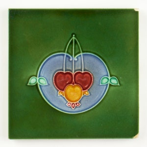 May include: A green ceramic tile with a blue and white circle design featuring three stylised cherries in red, yellow, and orange. The cherries are surrounded by green leaves.