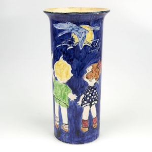 Pode incluir: Um vaso de cerâmica azul com um design caprichoso que apresenta duas crianças e um pássaro voando na frente de uma lua cheia. O vaso é decorado com tinta amarela, verde, vermelha e branca.