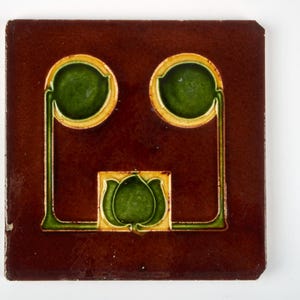 May include: Square brown tile with a decorative design. The tile features two green circles with yellow borders and a green floral shape at the bottom. Green lines connect the circles and the floral shape. The tile has a glossy finish.