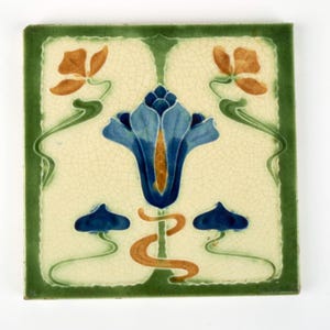 May include: Square ceramic tile with a green border and a cream-colored background. The tile features a blue flower with orange and green accents, and two orange flowers. The design is in an Art Nouveau style.