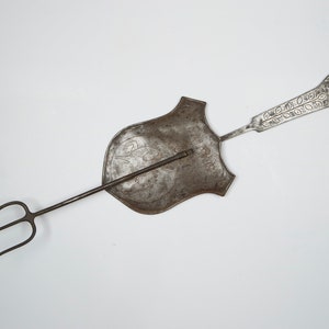 Puede incluir: Una herramienta de chimenea vintage de metal con un tenedor de tres puntas y un mango en forma de escudo. El mango tiene un dise&ntilde;o ornamentado y un bucle en la parte superior.