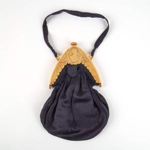 May include: A vintage black satin purse with a decorative frame. The frame is a light tan color with a cameo design and a dark handle. The purse has a gathered bottom and a black satin handle.