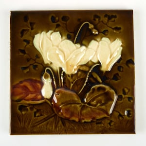 May include: Square ceramic tile with a floral design. The tile features cream-colored flowers, brown leaves, and a dark brown background. The flowers are the focal point, with detailed petals and stems.