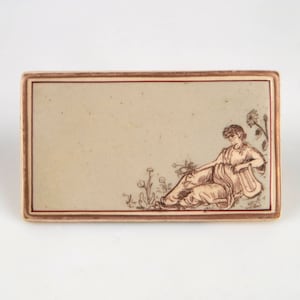 May include: A rectangular ceramic tile with a brown border and a cream background. The tile features a brown line drawing of a woman sitting in a field of flowers, holding a musical instrument.