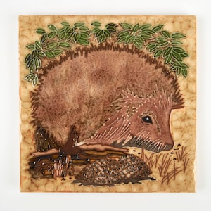 May include: A ceramic tile with a brown and white hedgehog design. The hedgehog is surrounded by green leaves and is curled up on a brown log.