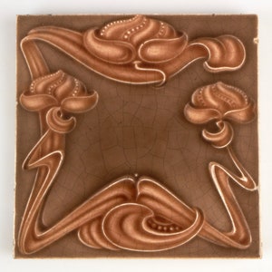 May include: A brown ceramic tile with a decorative design of stylized flowers in a light brown glaze. The tile has a crackled glaze finish.