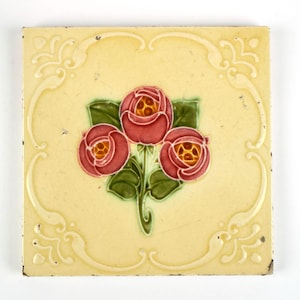 May include: A square ceramic tile with a cream-colored background featuring a central embossed design of three pink roses with green leaves. The tile is framed by a raised, swirling border pattern in a matching cream color, with visible surface wear.