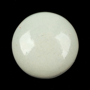 Puede incluir: Un objeto cerámico redondo de color celadón pálido con un acabado brillante. La superficie tiene un sutil patrón de craquelado y está sobre un fondo negro. El objeto parece ser una tapa o una pieza decorativa.