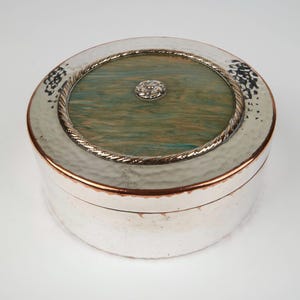 May include: A round, silver-toned box with a hammered texture. The lid features a central section with green and brown marbling, framed by a silver rope design and a decorative silver knob. The rim has a copper-colored band.