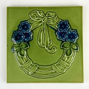 May include: A green ceramic tile with a raised design of a wreath of blue flowers and a green bow. The wreath is made of woven branches.