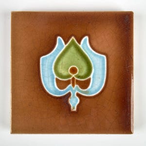 May include: Square brown ceramic tile with a central floral design. The design features a green, blue, and white stylized flower. The tile has a crackled surface texture.