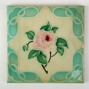 May include: Square ceramic tile with a floral design. The tile features a central image of a pink rose with green leaves and black stems. The border is a light teal color with a decorative swirl pattern.