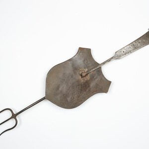 Puede incluir: Una herramienta de metal vintage con una cabeza en forma de escudo y un mango largo. El mango tiene una horquilla de tres puntas en un extremo y un anillo en el otro.