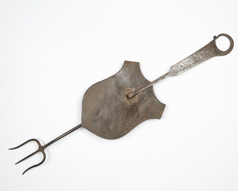 Puede incluir: Una herramienta de metal vintage con una cabeza en forma de escudo y un mango largo. El mango tiene una horquilla de tres puntas en un extremo y un anillo en el otro.