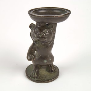 Può includere: Statuetta in bronzo di una creatura simile a un orso che tiene un piccolo piatto rotondo sopra la testa. La statuetta ha un volto dettagliato e poggia su una base circolare. Il design suggerisce un oggetto decorativo o funzionale.