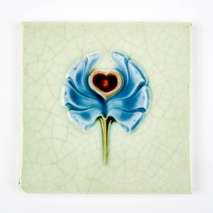 May include: Square ceramic tile with a light green background and a blue floral design. The flower has a heart-shaped center in a darker shade of red. The tile has a cracked, aged appearance.