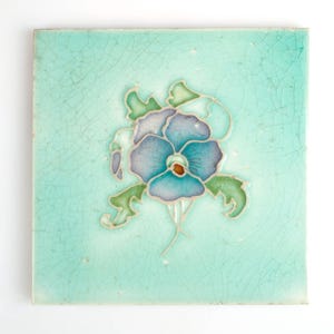 May include: A square ceramic tile with a light blue background and a stylized blue and purple flower with green leaves.