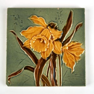 May include: A square ceramic tile featuring a raised relief design of two yellow daffodils with brown stems and leaves against a muted sage green background. The floral motif is centred on the tile, showcasing detailed petals and trumpet centres.