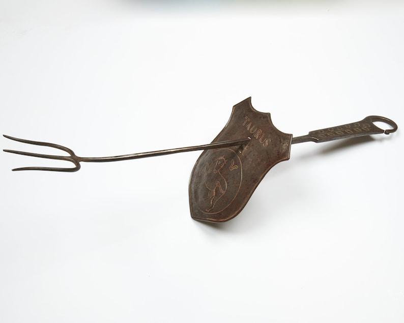 Puede incluir: Un utensilio de cocina de metal vintage con un mango largo y un tenedor de tres puntas. El utensilio tiene una cabeza en forma de escudo con la palabra "Taurus" y un dise&ntilde;o de toro estilizado.