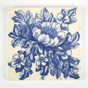 May include: A square ceramic tile featuring a central blue and white floral pattern. The design displays a large, detailed flower surrounded by smaller blossoms and intricate, lobed leaves, all rendered in a deep blue pigment on a cream background.