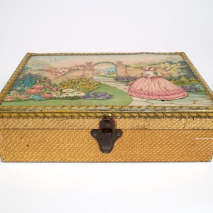 Peut inclure: Une boîte de rangement rectangulaire en métal avec un couvercle à charnière orné d'une illustration vintage représentant une personne en robe rose dans un jardin avec une arche en pierre. Les côtés ont un motif texturé jaune et marron.