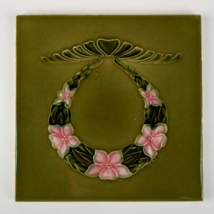Può includere: Piastrella quadrata in ceramica con sfondo verde e design a ghirlanda. La ghirlanda è composta da foglie verde scuro e fiori rosa. La piastrella ha una finitura lucida, probabilmente per uso decorativo.
