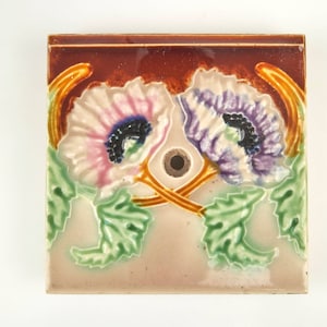 Może przedstawiać: Płytka ceramiczna z kwiatowym wzorem. Płytka jest brązowa, biała, różowa, fioletowa i zielona. Kwiaty są stylizowane i mają vintage'owy wygląd. Płytka ma otwór w środku.