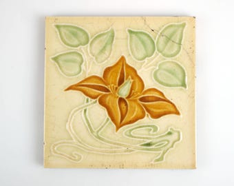 May include: A ceramic tile with a floral design. The tile features a large orange flower with green leaves and a cream background.