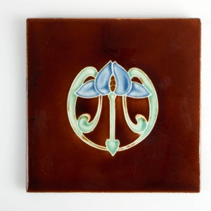 May include: A square ceramic tile with a brown background and a stylised blue flower design in a green circle.