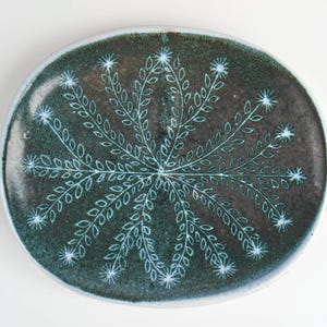 May include: A dark green oval ceramic plate with a white floral design. The design features a stylized flower with leaves radiating outward from the center.