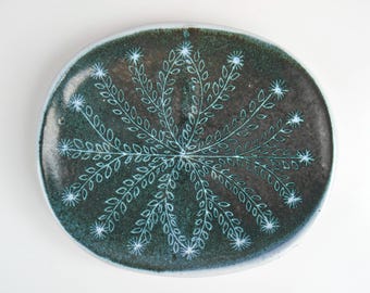 May include: An oval ceramic plate in a dark green colour with a white floral design. The design features a stylised flower with leaves radiating outwards from the centre.