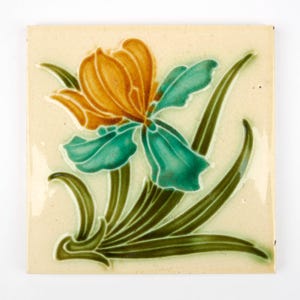 May include: Square ceramic tile with a floral iris design. The flower has orange petals and teal accents, with green leaves and stems. The tile has a cream background and is suitable for decorative use, such as a kitchen or bathroom.