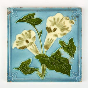 May include: Square ceramic tile with a light blue background and a floral design. The design features white flowers with brown accents and green leaves. The tile has a decorative border and is likely vintage.