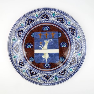 Könnte beinhalten: Ein dekorativer Keramikteller mit detailliertem blau-weißem Blumenrand. Die Mitte zeigt ein blaues Wappen mit einem weißen Hirsch und goldenen Akzenten auf braunem Hintergrund. Das Design des Tellers ist aufwendig und kunstvoll.