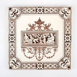 Könnte beinhalten: Eine braune und weiße Keramikfliese mit einem dekorativen Design, das eine zentrale Urne mit einem floralen Motiv und Figuren in einer Gartenlandschaft zeigt. Die Fliese ist von einem Rand aus komplizierten Schnörkeln umgeben.