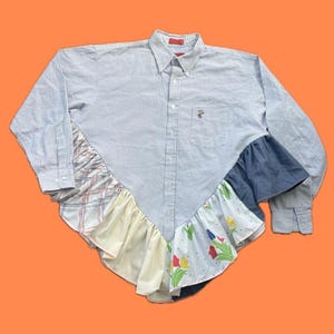 May include: A long-sleeve button-down shirt featuring a light blue and white pinstripe pattern. The lower section is accented with a V-shaped hemline composed of layered, ruffled fabric panels in solid yellow, white with a floral print, and navy blue.