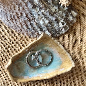May include: A small, handmade ceramic dish with a turquoise and tan color scheme, holding two silver rings. The dish is placed on a woven, tan surface, with a large seashell and starfish in the background.